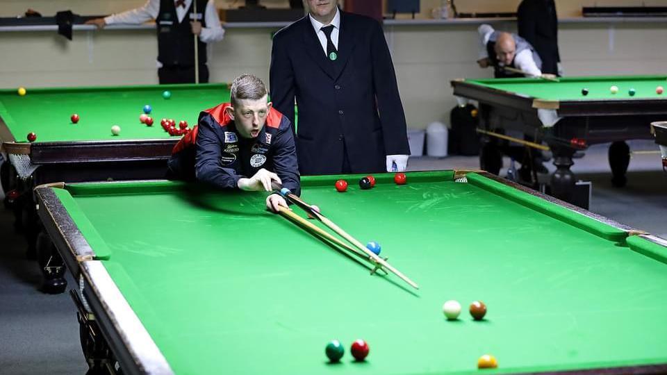 David Church wearing a black and red waistcoat leaning over a snooker table and taking a shot while using a rest. A referee is standing behind him. Play is under way at two other snooker tables, which can be seen behind Mr Church.