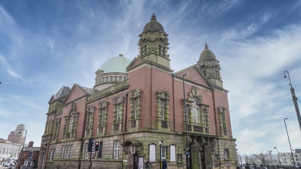 The outside of the building. The building has a green dome and two towers. Traffic lights and street lamps and also in the picture. 