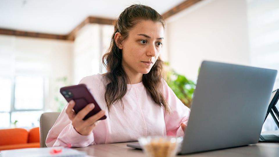 A stock image of a woman with long brown hair and a pink jumper, sitting a desk with a laptop on a desk and a mobile phone in her hand