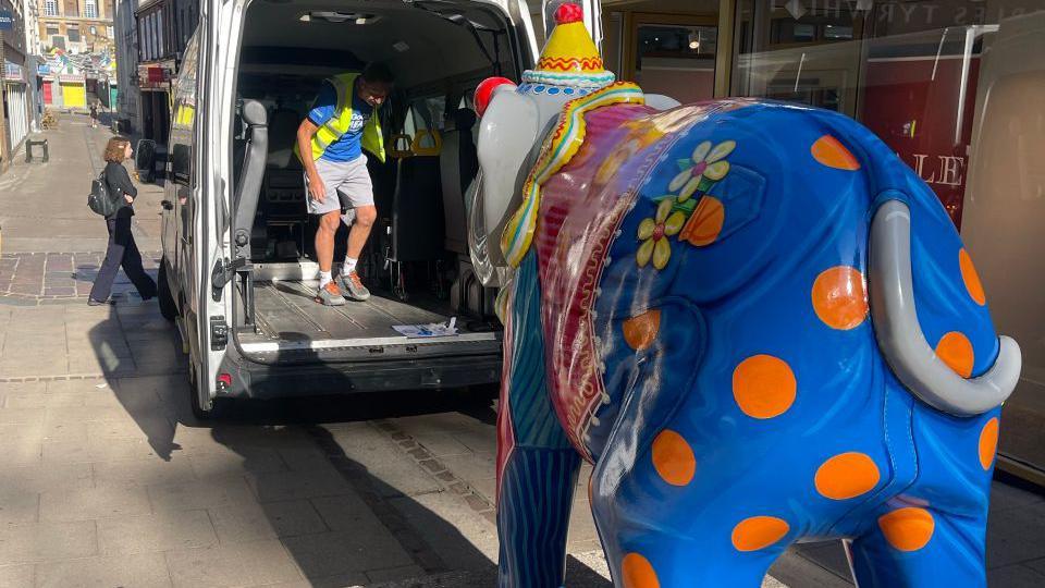 A statue of a elephant, which is painted blue and orange, is standing on a concrete walk way. it is being loaded into a van as it needs a repair. 