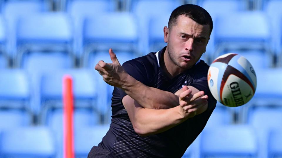 Charlie Chapman spins a pass out of his left hand sending the ball straight at the viewer. He watches it closely as it flies away from him. In the background there are empty light blue seats which are blurred and the top part of a red training stick.