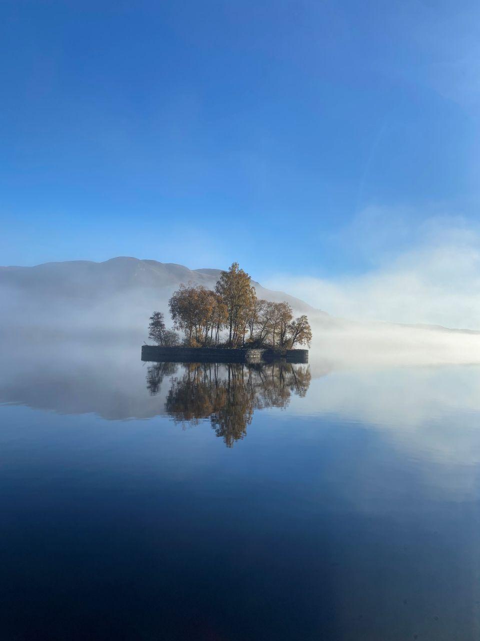 A small island with trees set on a lake. The sky is blue and a single cloud is reflected in the water