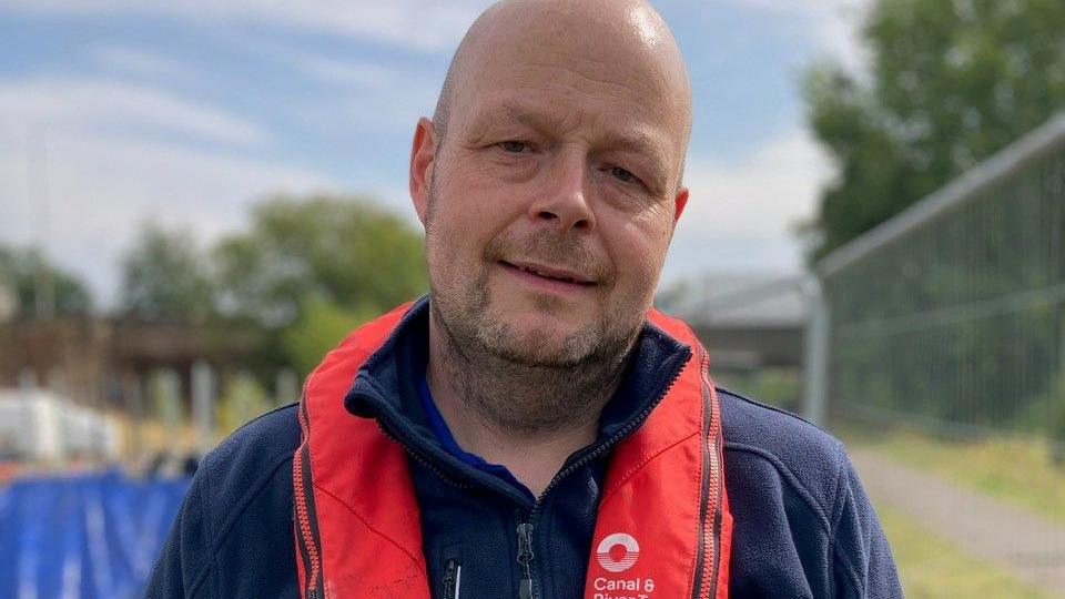 A man wearing a blue fleece and CRT life jacket. He is looking at the camera in front of a blurred background of the canal and fencing.