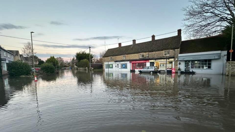 A flooded residential road along a parade of shops, with the water flowing all the way down the road 