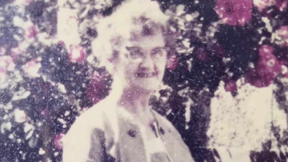An old photo of Nancy Barber, who has short curly hair and is wearing glasses, against a floral backdrop.