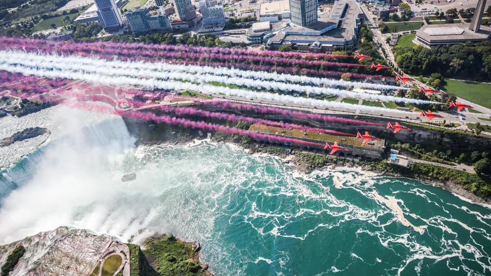 The nine Red Arrows planes flying in formation, there are red and white streams behind them as they fly over the waterfall. The photo is taken from above the planes.