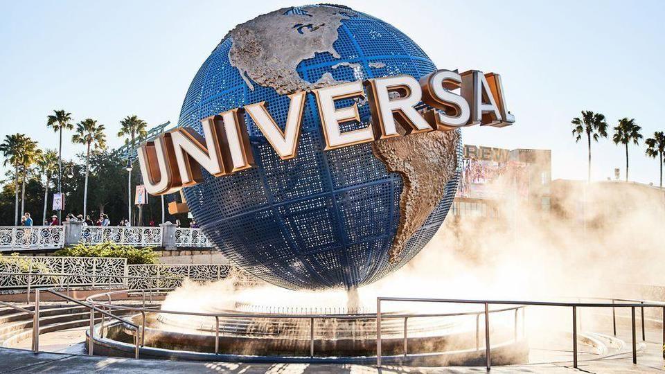 The Universal Studios globe is in a theme park, it is situated on top of a water fountain and in the background there are palm trees.