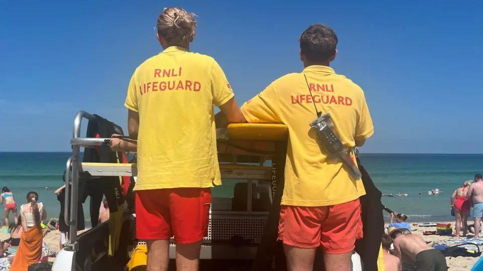 Two lifeguards, wearing yellow tops which said 'RNLI Lifeguard' and red shorts. There are looking out to the water and facing away from the camera. There are people on the beach facing the sea. In the background is the blue sea and light blue sky.