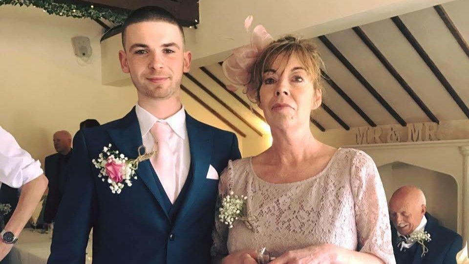 Liam Harvey at a wedding prior to his diagnosis, wearing a pale pink shirt and tie underneath a navy blue blazer pinned with a pink corsage. He is standing beside his mother Alex, who is wearing a pink lace dress and fascinator. 