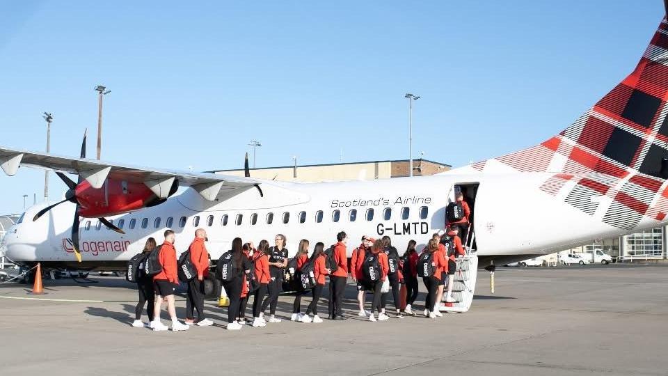 Members of the Isle of Man team wearing red jackets boarding a Loganair plane at Ronaldsway Airport.