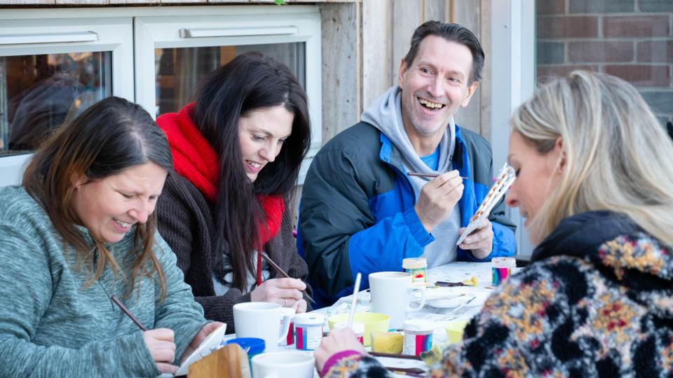 Three women and a man smile as they sit around a table making crafts. They each have a hot drink in front of them.