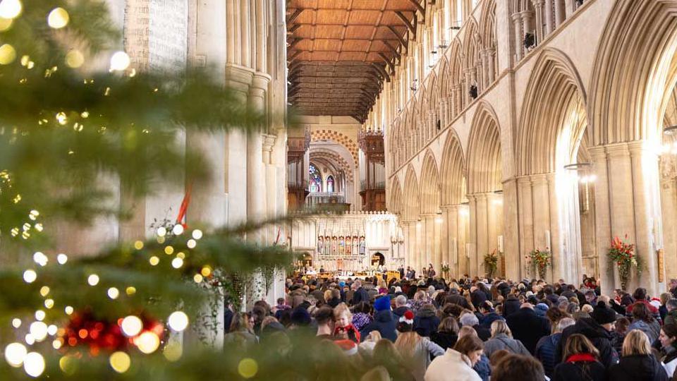 The nave of St Albans Cathedral is filled with members of the public listening to a Carols on the Hour service. There is a Christmas tree in the foreground.