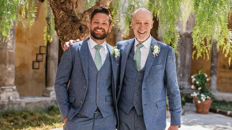 A young man and an older man wear matching blue suits and mint green ties, with corsages on their suits. They grin at the camera, with the older man's hand on the younger man's shoulder.