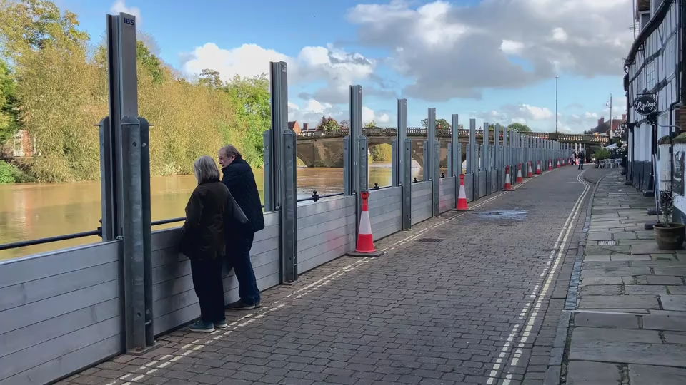 A partially-erected metal flood barrier along a brick-cobbled street. It consists of strips of metal fencing placed between upright posts. A couple is looking over the barrier at the rising river.  