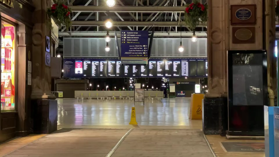 The concourse at Glasgow Central Station. It is empty. A yellow wet floor sign is in the middle. There is a shiny white floor. In the background are screens displaying travel information.