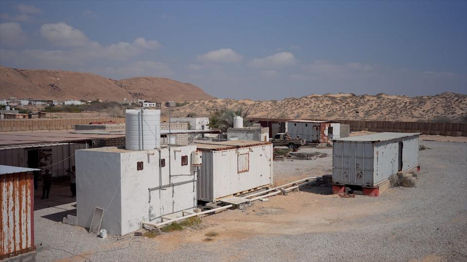 One white shipping container and two grey ones are seen on a gravel area with sandy hills behind them. A small white building with a water tank on it is next to one of them.