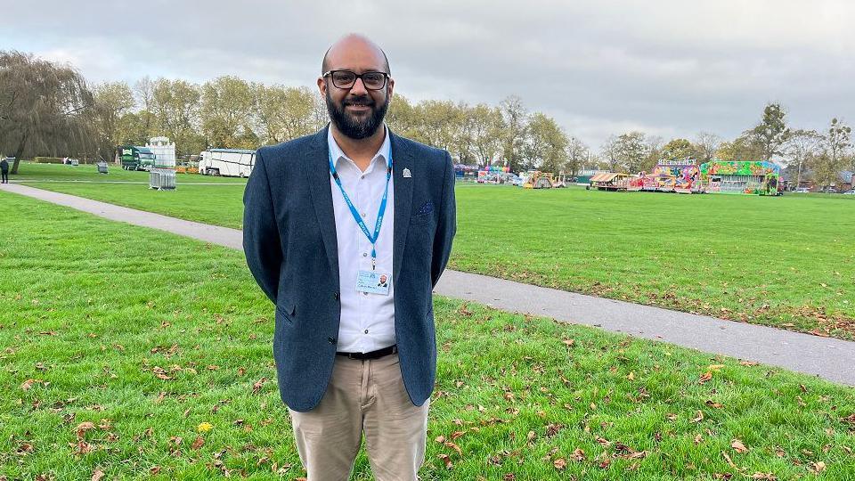 Local councillor Bal Anota standing at the Walks, a public park in King's Lynn. He is wearing a blue jacket, white top and tan chinos. 