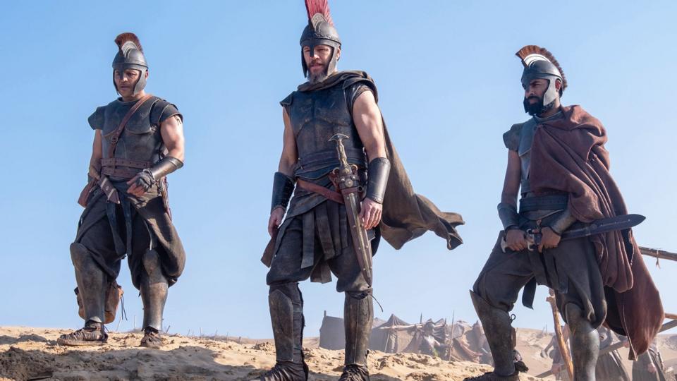 Three soldiers from the film The Odyssey standing in a row looking into the distance with a blue sky behind them