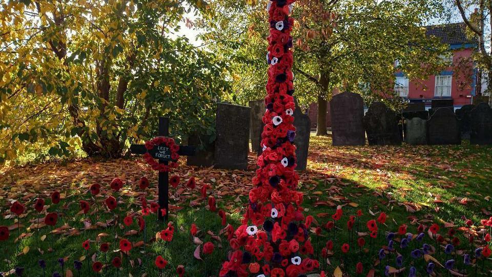 A graveyard in autumn with pops of colour from knitted poppies on the ground and placed up a tree's trunk