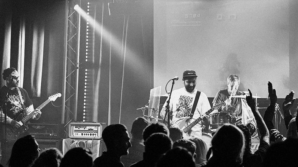 Black and white photo of a band on stage. One of the men is wearing a cap and a white T-shirt and holding a guitar. 