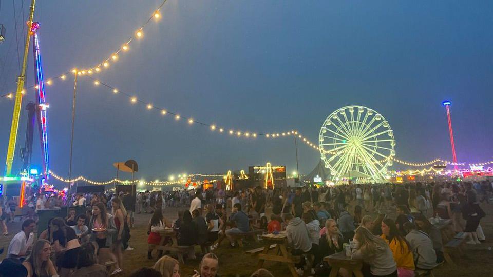 A night time shot showing groups of people at Boardmasters sitting at tables enjoying drinks and food, with a lit-up Ferris wheel in the background and other bright lights