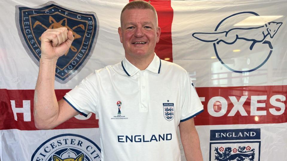 Darren Thompson smiling with his hand in a fist and standing in front of an England flag, which has four badges in each quarter.
