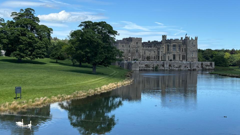 A view of Raby Castle near Staindrop in County Durham with two swans swimming up the lake towards it. The sky is a bright blue and sun is lighting up trees on the far side of the bank.