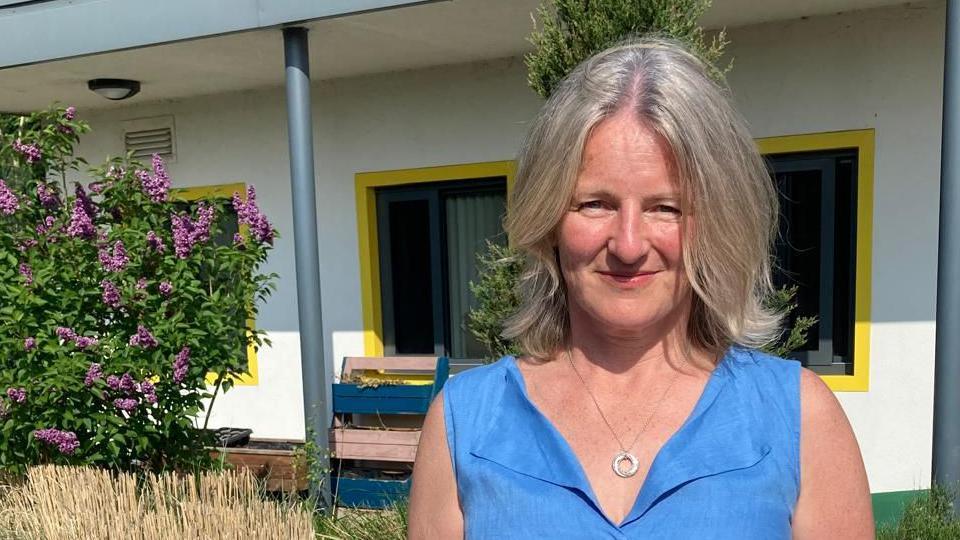 Dr Black stands outside a modern building, which is painted white with bright yellow colours surrounding the window frames. She is wearing a bright blue top and has medium length greying hair. Dr Joe is looking at the camera, with a hoop pendant hanging on a necklace. Surrounding her are bright bushes, plants and flowers, in a sunny garden area.