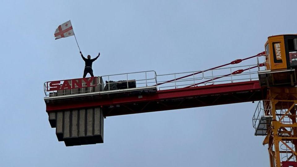 A Georgian flag is waved from a crane at protesters passing underneath