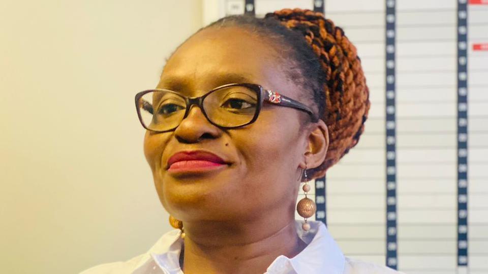 Sibongile Tshabalala, wearing a white blouse, lipstick, glasses and dangly earring with her hair in a bun, smiles.