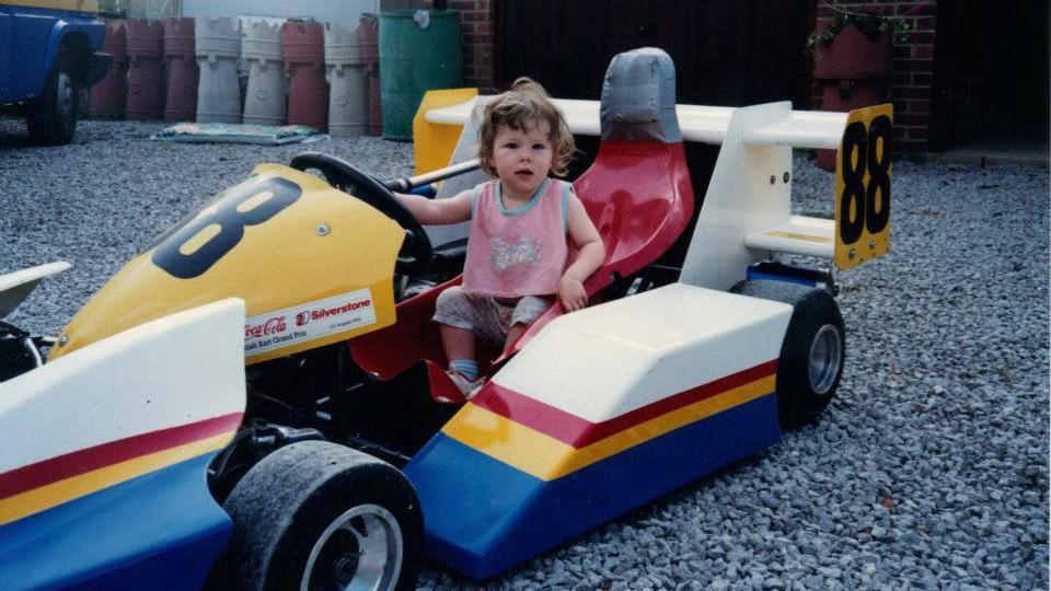 A family snap of Abbie Eaton as a young girl sitting in a racing car. The colourful, single-seat car has yellow, white, red and blue stripes, and a red seat. The number 88 is printed on the front and rear spoiler.