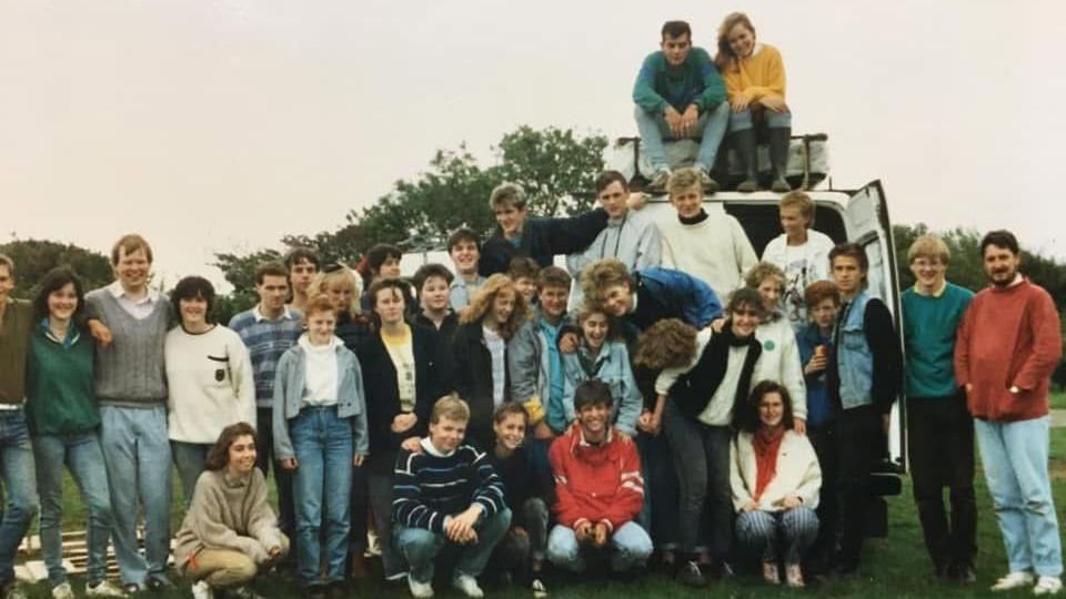 A group of students in 1985. Two are sitting on top of a white van with its doors open. Some are standing up, others are crouching down, making up four rows of students, all outside, standing around a white van. 