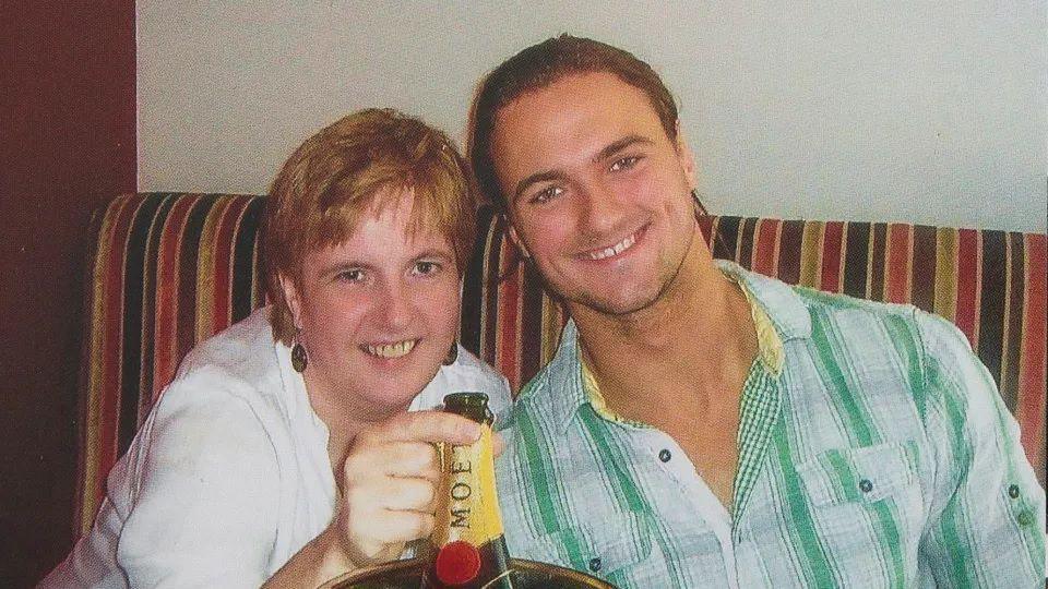 Angela and Drew (as a young man) smile for the camera, with a bottle of champagne sitting in front of them. She is in a white blouse and he is in a green and white shirt. They are sitting on a brown, red, yellow and green striped sofa.