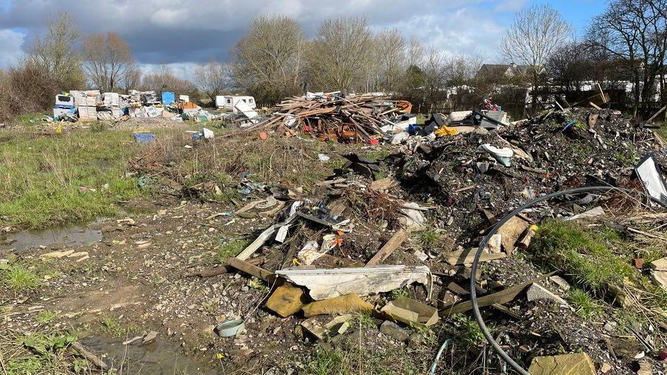 A grassland which is covered in mud, debris, a bonfire and in the distance, piles of pallets and abandoned caravans