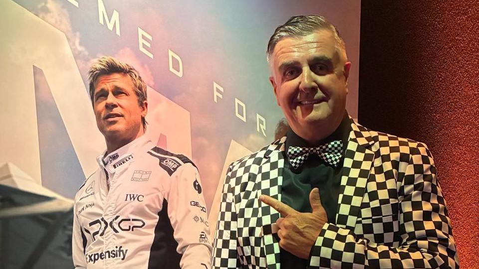 Martin Donnelly is standing on the right looking towards the camera smiling He is wearing a black and white chequered suit jacket, and a black shirt with a tie to match. He is point to a poster of the film which has a picture of Brad Pitt wearing a white racing suit.