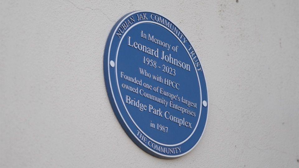 A blue plaque on a white wall that reads: "In Memory of Leonard Johnson 1958 to 2023, who with the HPCC founded one of Europe's largest owned Community Enterprises, Bridge Park complex in 1987"