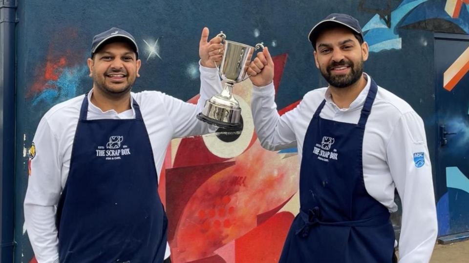 Two owners of a fish and chip shop stand outside it holding the large silver trophy.
