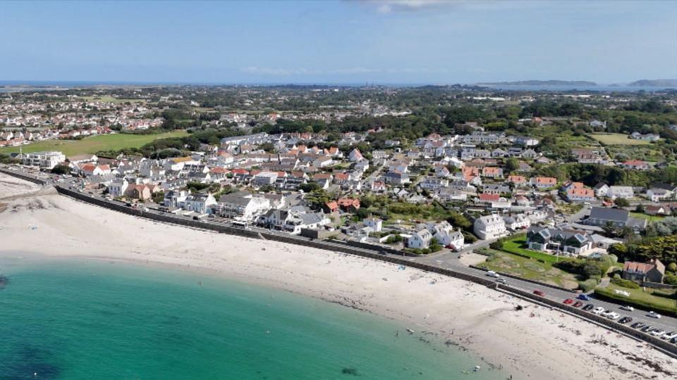 An aerial view of Guernsey from above, there is blue sea and white sand and then housing and buildings behind in the distance. The sky is blue.