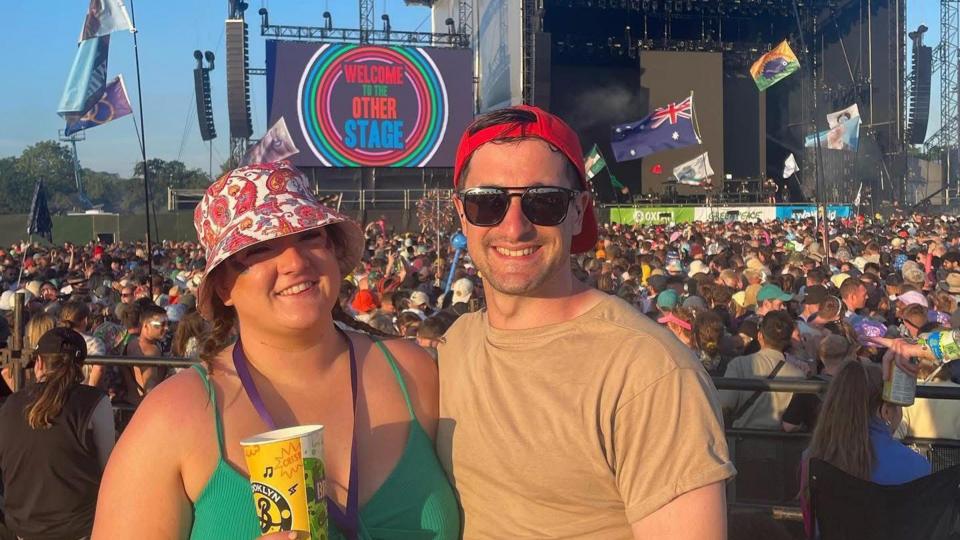 A woman and a man stand illuminated by sun in front of a festival stage, with a large crowd watching the stage.