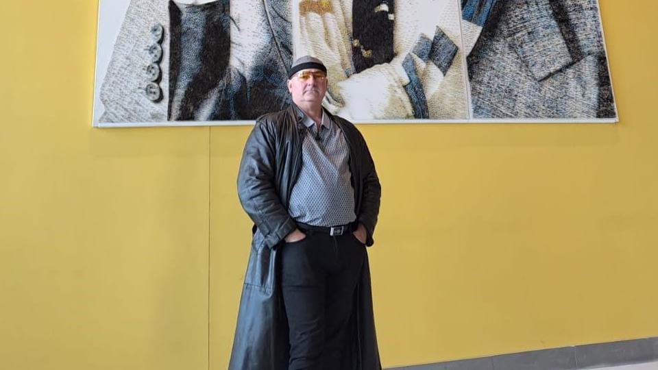 Artist Marcus Levine stands in front of his artwork and a yellow wall. He is wearing a black leather coat and tinted glasses