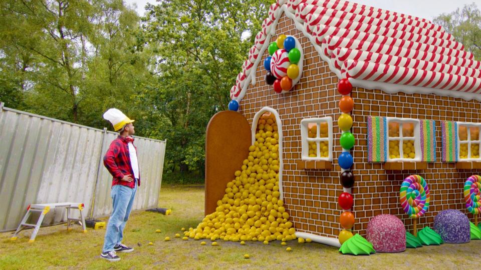 giant gingerbread house with gold nuggets spilling out the front door. a person with a chef hat stands with is hands on his hips, looking at the house