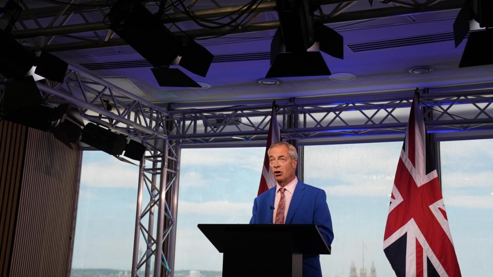 Nigel Farage is wearing a bright blue coloured suit jacket, white shirt and patterned tie. He is stood in front of a Union flag