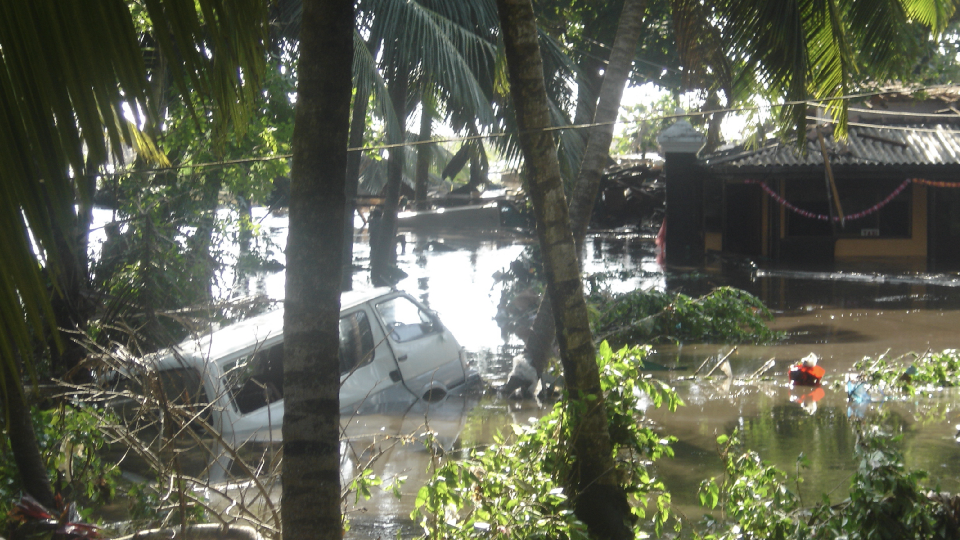 A white van is partly submerged in water, surrounded by palm trees and flooded huts