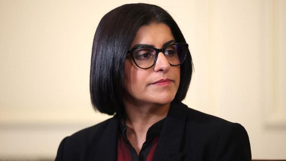 Home Secretary Shabana Mahmood is pictured looking off camera. She is wearing a dark jacket with a red and black top on beneath. 