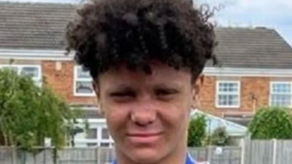 Harleigh has dark curly hair and is wearing a blue top. He stands in front of houses with a fence in front and trees behind them.