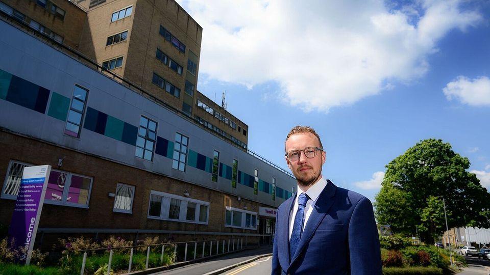 Man with glasses stood in front of a hospital. He's in a suit and tie.