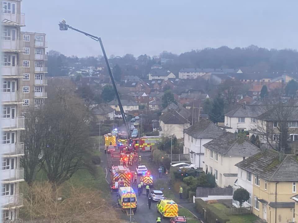 Half a dozen emergency service vehicles, including an aerial ladder, line the street outside the high-rise. Several houses with front gardens are located on the right