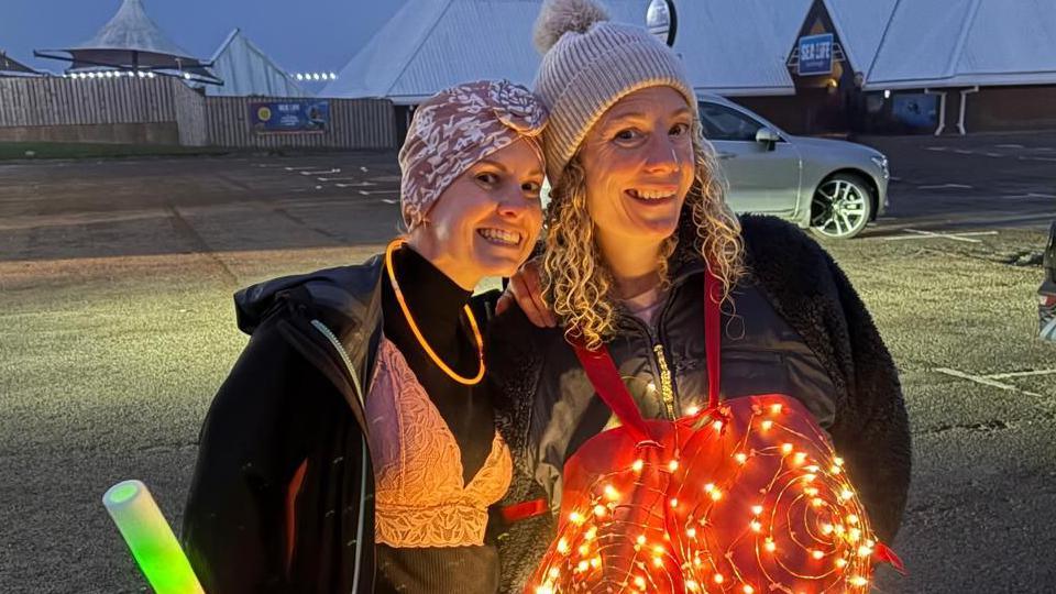 Two women stand in a car park at dusk. The woman on the left wears black clothing with a pink head covering and a pink bralette over her clothes and holds a large glowing stick emitting green light. The person on the right wears a white knit hat and a red costume lit with small string lights.