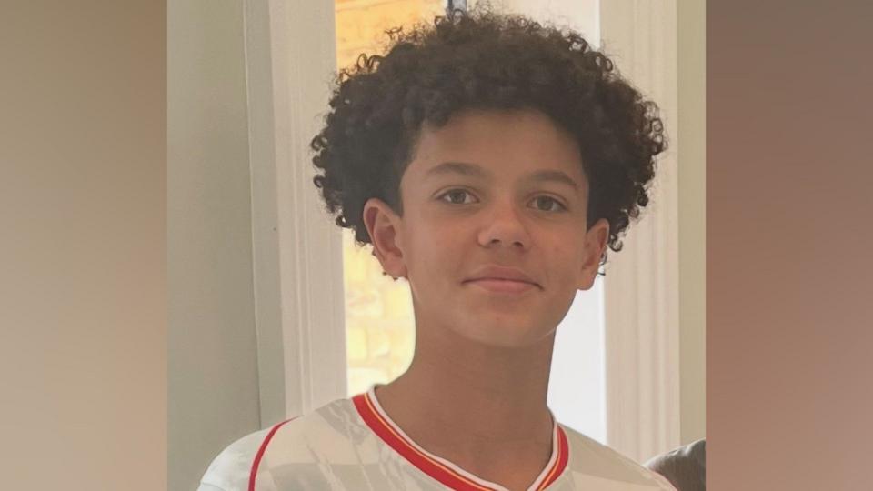 Noah smiling at the camera. He is wearing a white football shirt with red edging and has short curly hair.