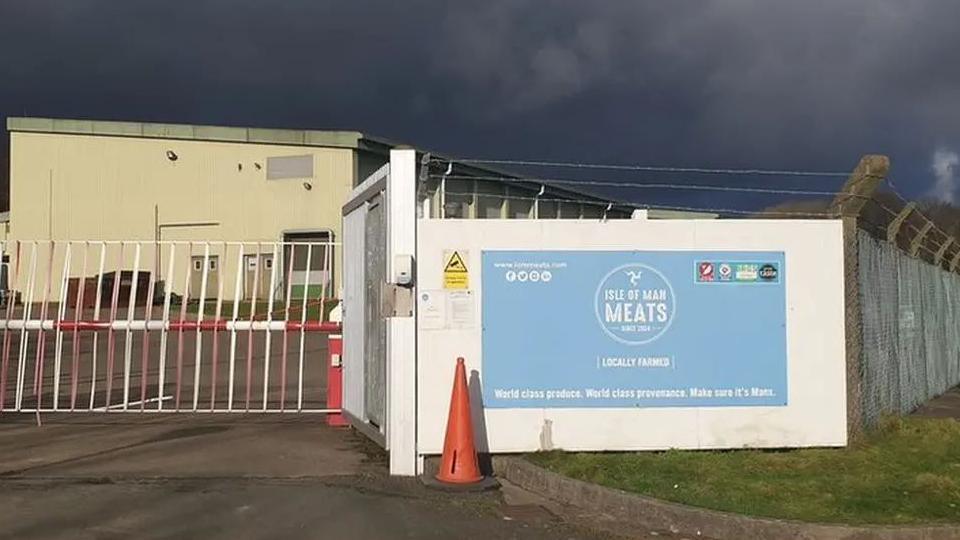 The exterior of a building which is protected by tall fences topped with barbed wire. A metal gate runs along the front entrance, which also has a large pale blue sign with the Isle of Man Meats logo on it.